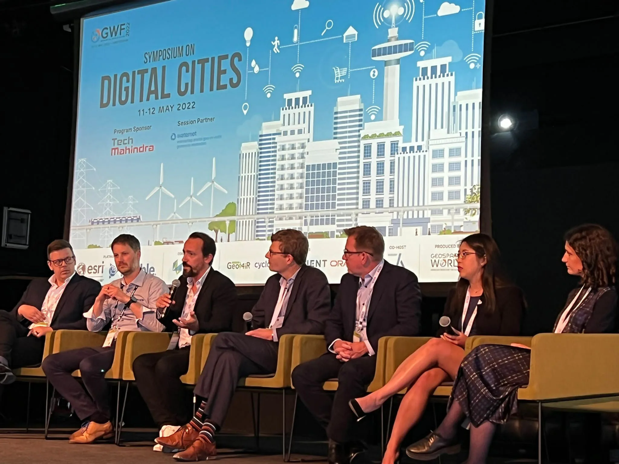 Panel discussion at the Symposium on Digital Cities (May 2022), with six speakers seated on stage in front of a large screen displaying a smart city illustration and event sponsors.