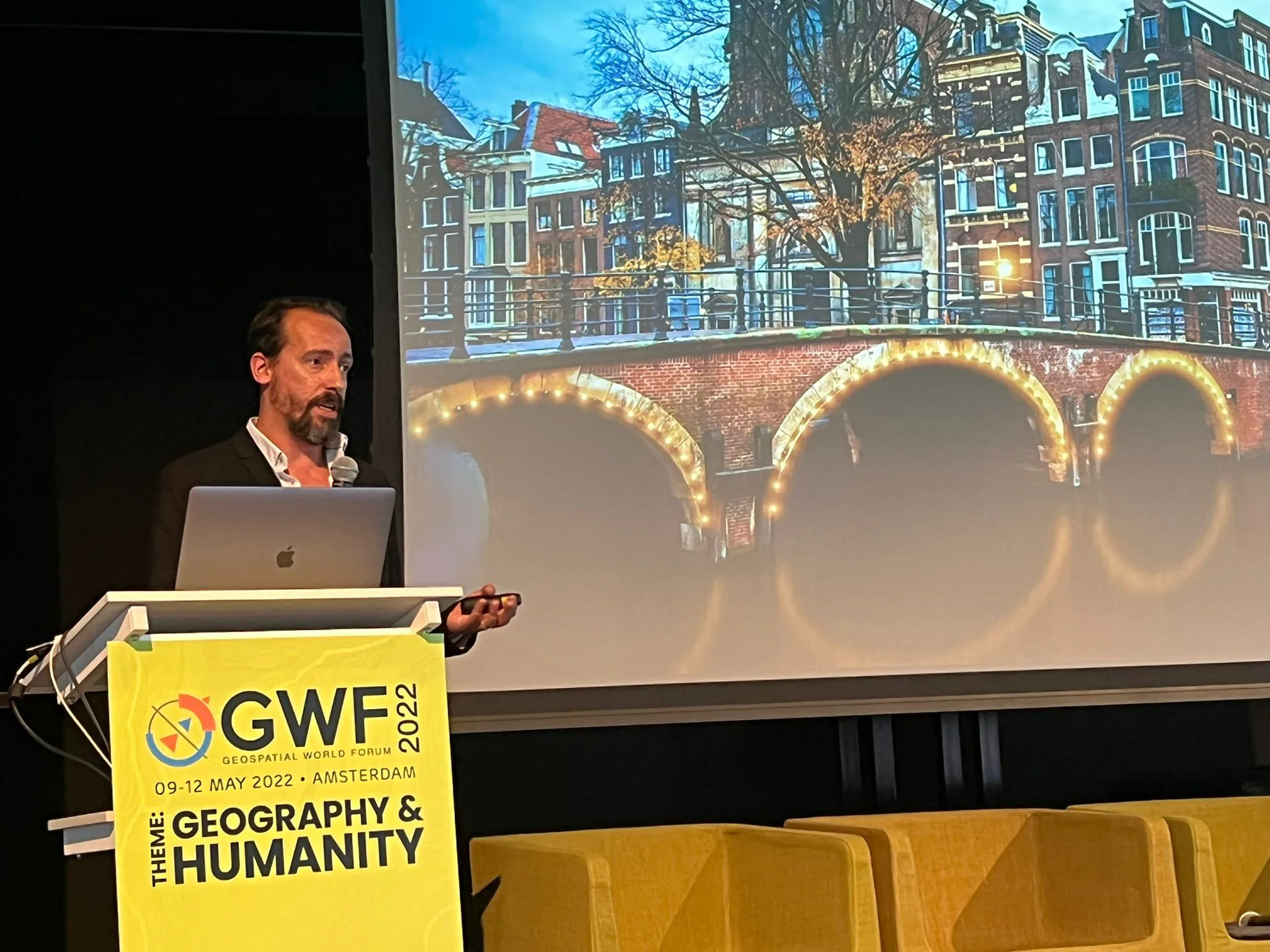 Speaker presenting at the Geospatial World Forum 2022 in Amsterdam, standing at a podium labeled “GWF 2022 – Geography &amp; Humanity,” with an image of Amsterdam’s canals projected behind him.