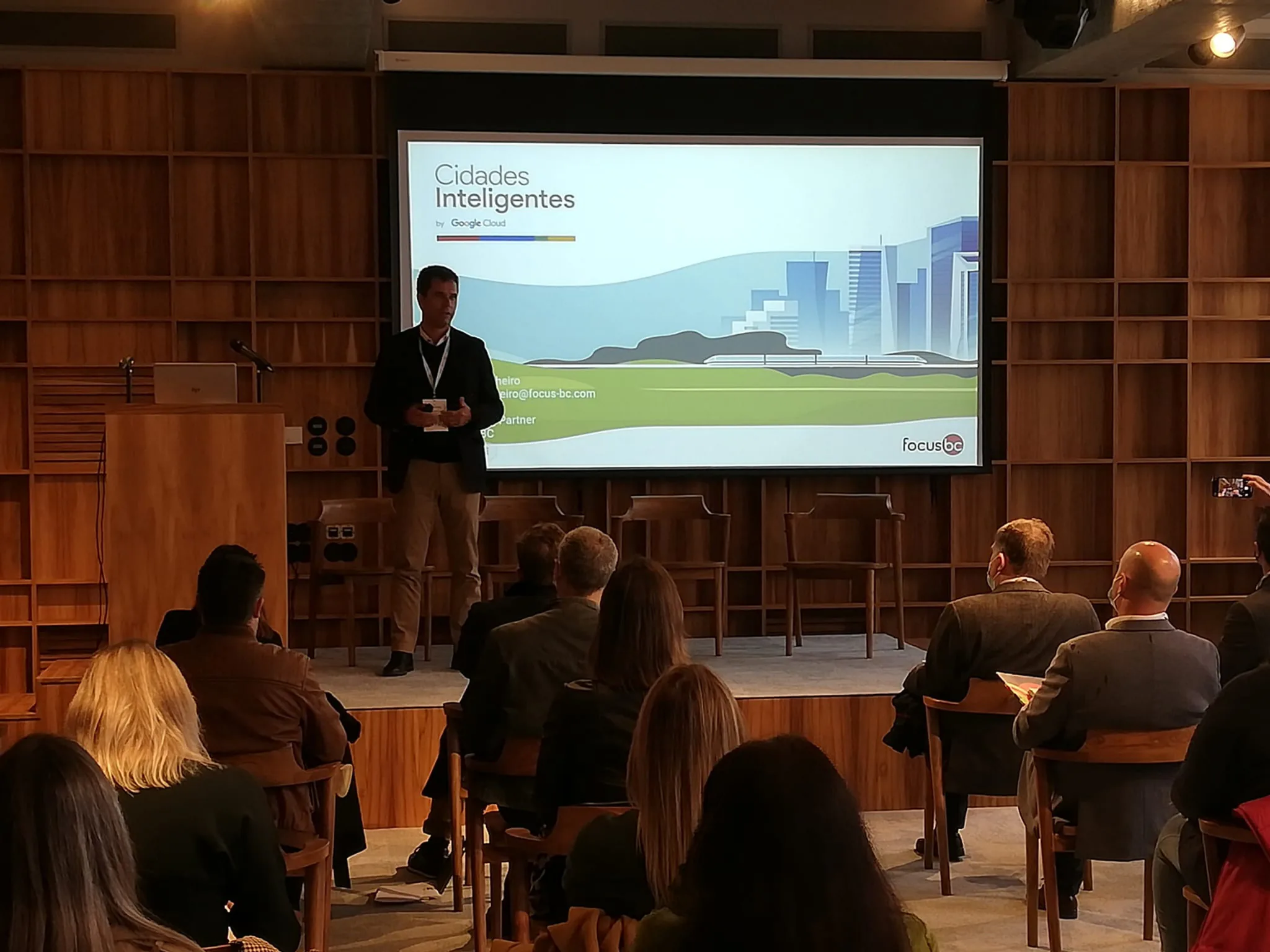 Speaker presenting “Cidades Inteligentes by Google Cloud” to an audience in a wood-paneled conference room, with the Focus BC logo on the slide.