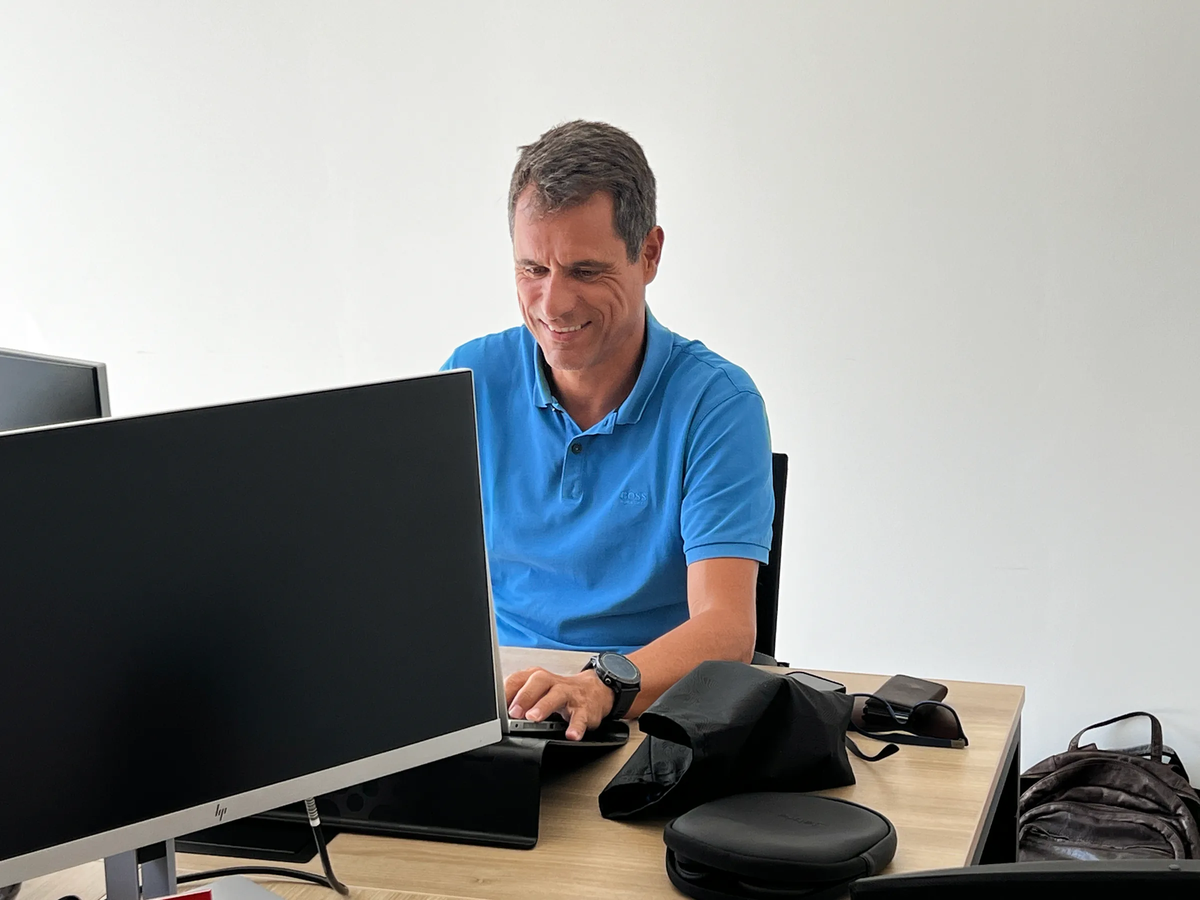 Vasco in a blue polo shirt sitting at a desk in a bright office, smiling while working on a desktop computer, with a backpack and small personal items placed on the table beside him.